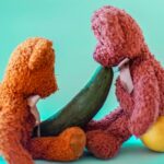 Two colorful teddy bears interacting creatively with fruits and cucumber.