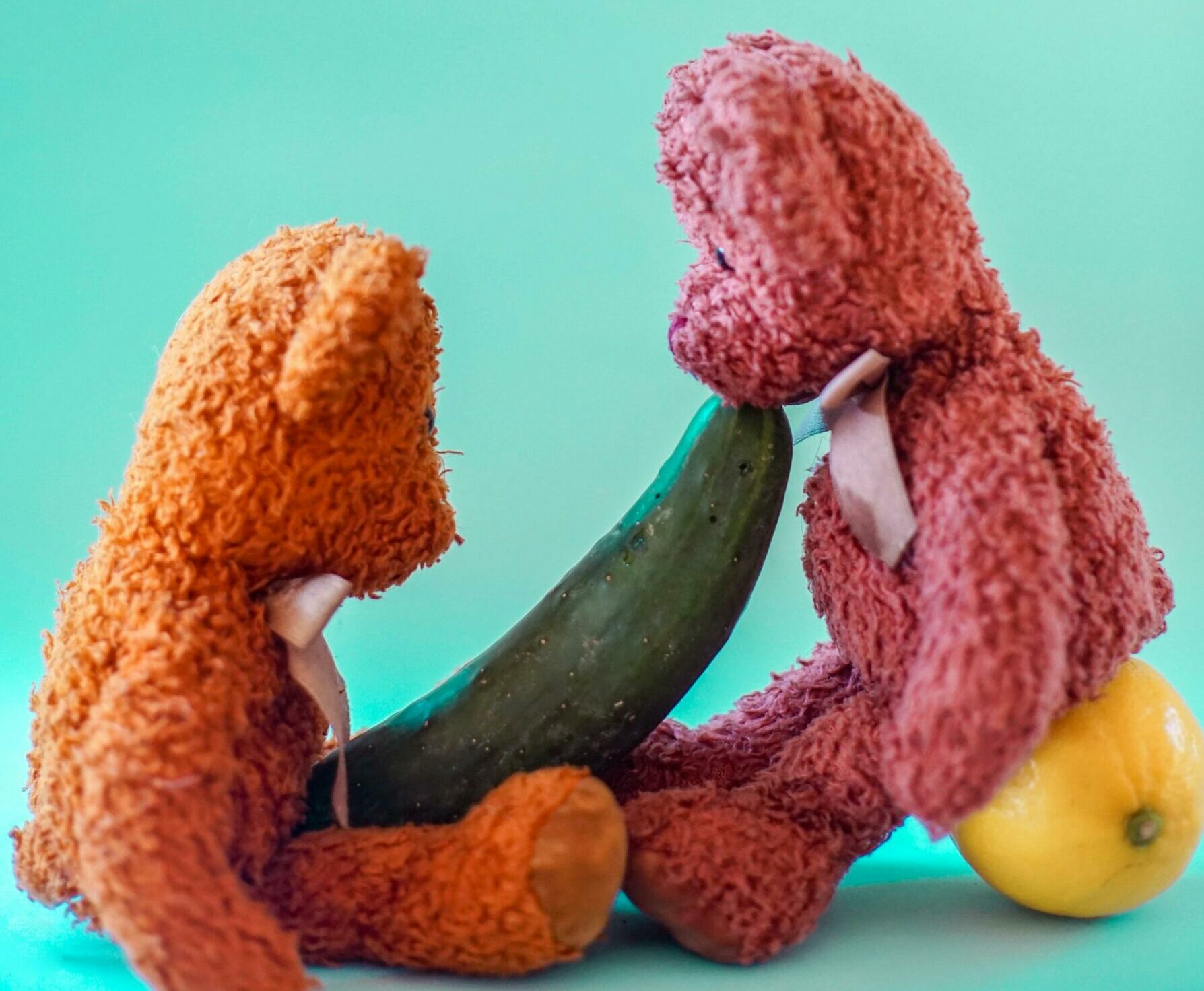 Two colorful teddy bears interacting creatively with fruits and cucumber.