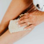 Close-up of a person exfoliating legs in the shower with a body scrubber.