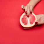 Close-up view of a hand with red nails holding a grapefruit against a red background. Symbolic and abstract.
