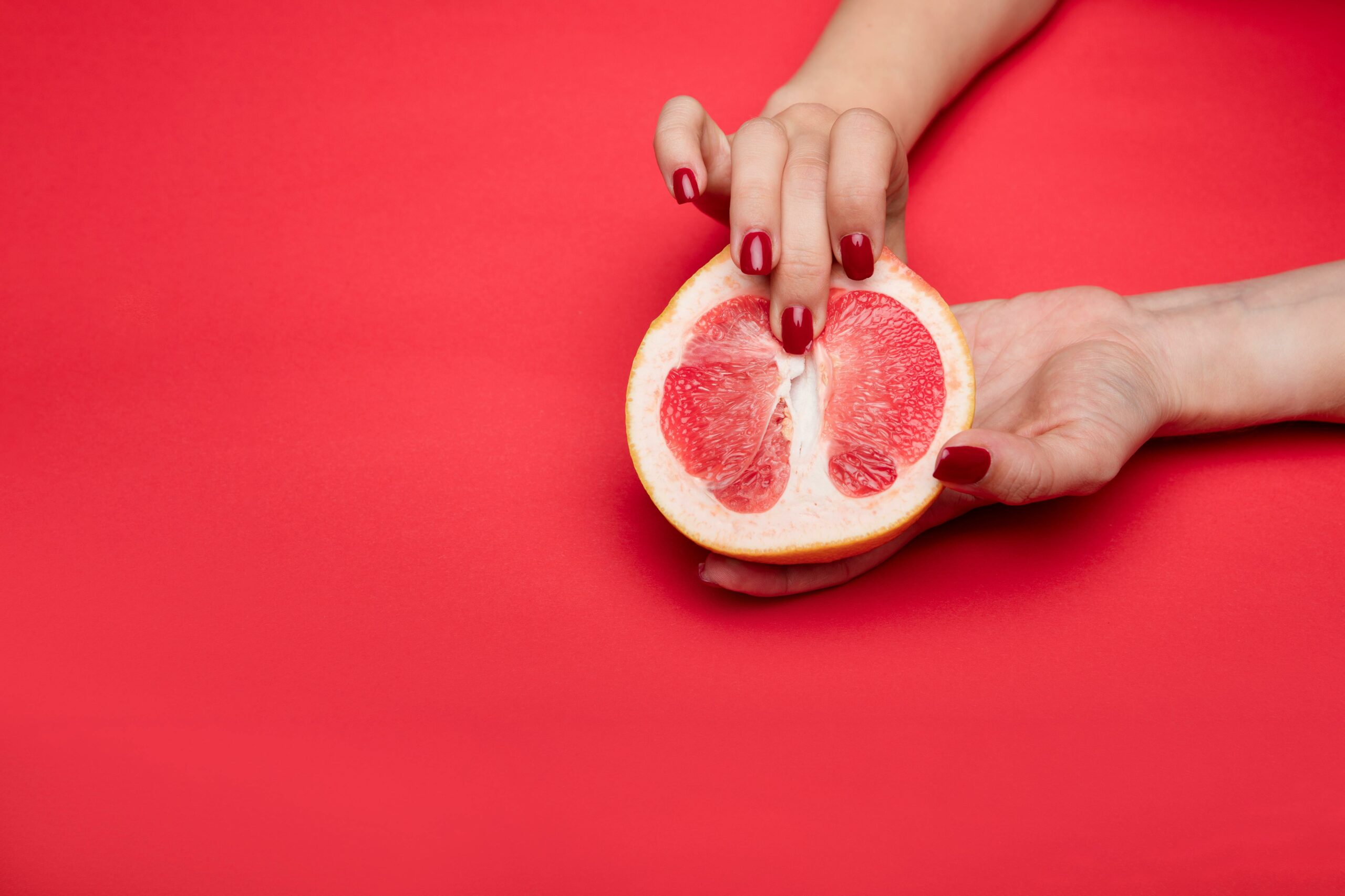 Close-up view of a hand with red nails holding a grapefruit against a red background. Symbolic and abstract.