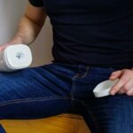 A man seated indoors holding an adult toy device, wearing jeans and a black shirt, with focus on the product.