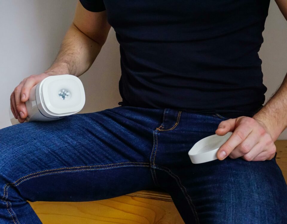 A man seated indoors holding an adult toy device, wearing jeans and a black shirt, with focus on the product.