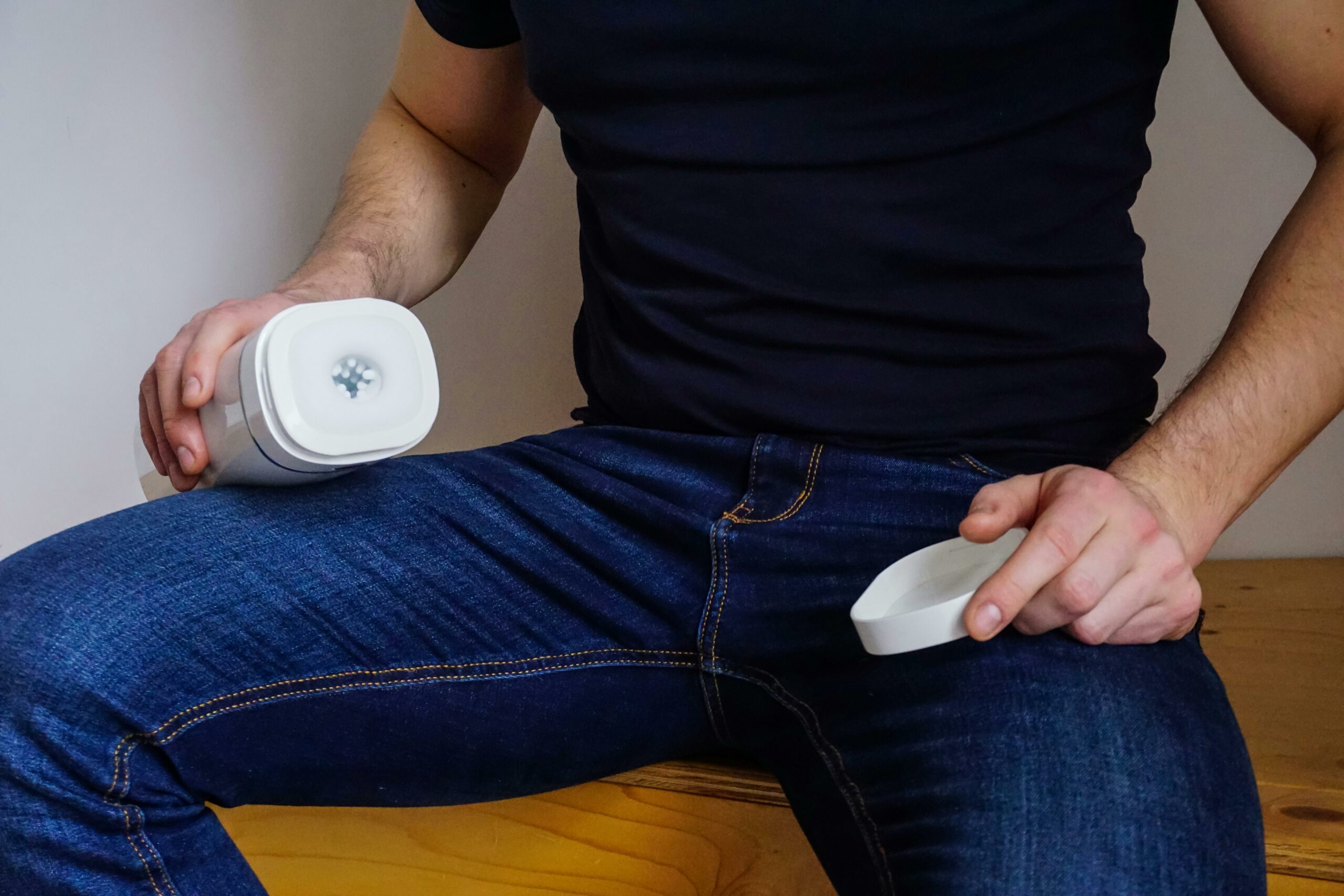 A man seated indoors holding an adult toy device, wearing jeans and a black shirt, with focus on the product.