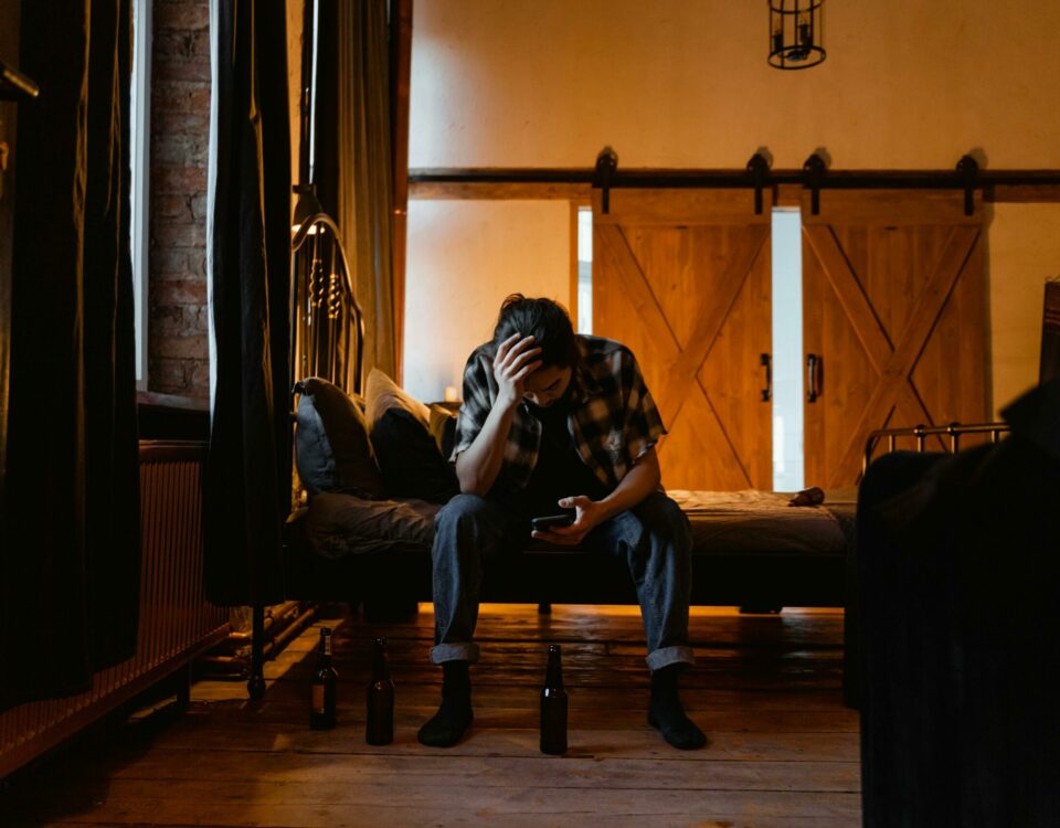 A man sits on a bed in a dark room, appearing pensive and stressed.