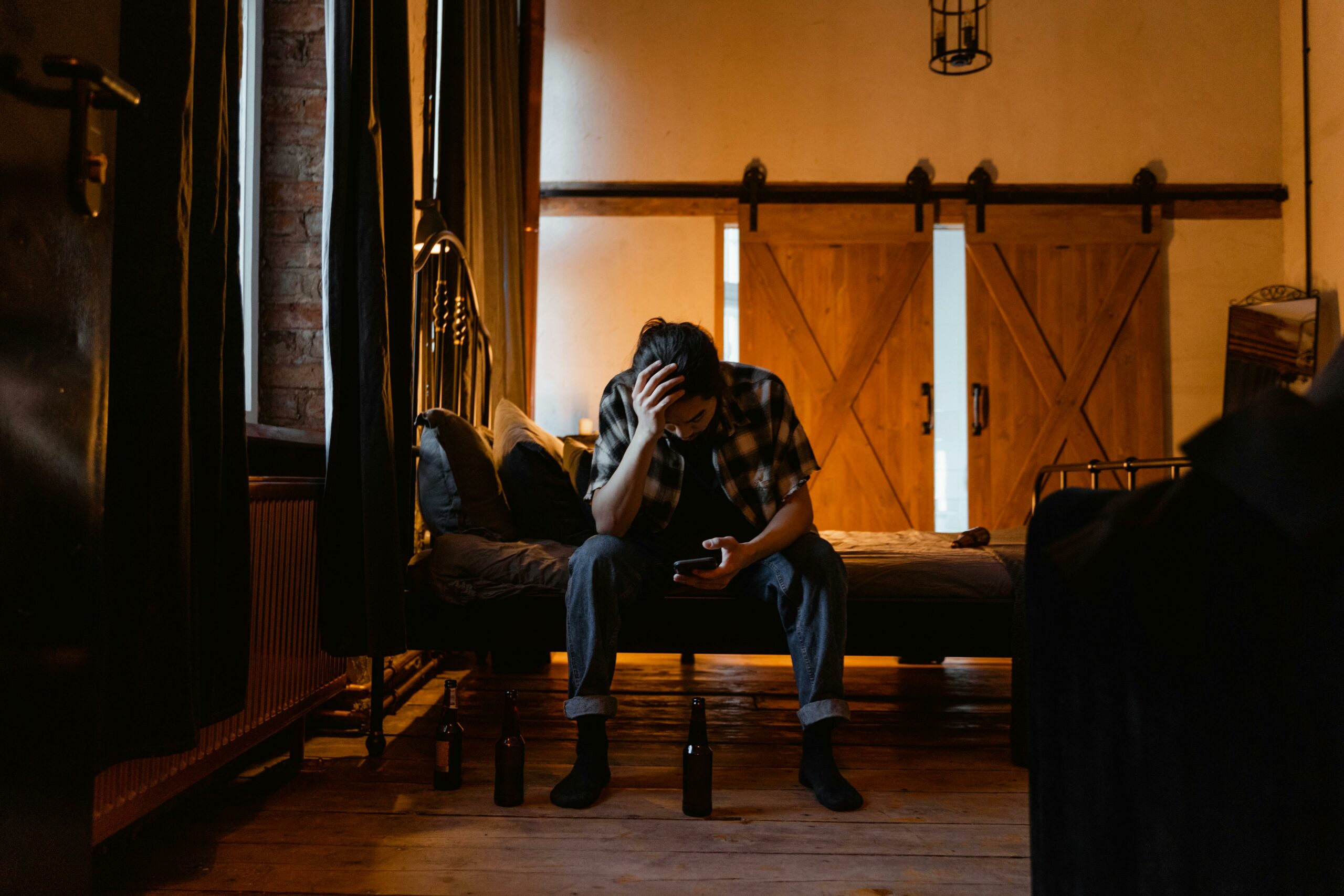 A man sits on a bed in a dark room, appearing pensive and stressed.