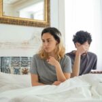A couple sits together in a bedroom, experiencing a moment of misunderstanding.